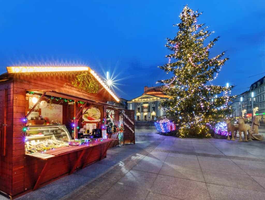 A wooden market stall with food stands beside a large decorated Christmas tree with colorful lights in a city square at dusk, capturing the enchanting spirit of the Best Christmas Markets in Poland.