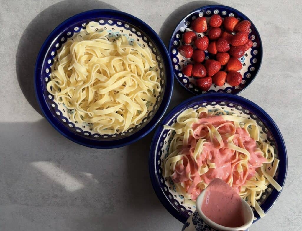 Three ceramic dishes feature plain fettuccine, fettuccine with pink sauce, and fresh strawberries—perfect for trying a Polish pasta favorite, Makaron z truskawkami. A small jug with extra pink sauce completes this strawberries recipe.