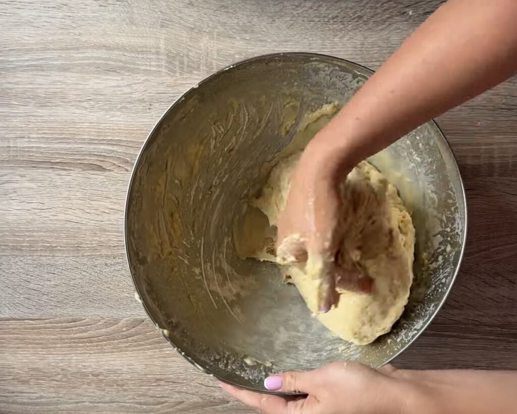 A person kneads dough by hand in a large mixing bowl on a wooden surface, preparing Polish Apple Yeast Buns (Drożdż&oacute;wki) using an easy recipe.