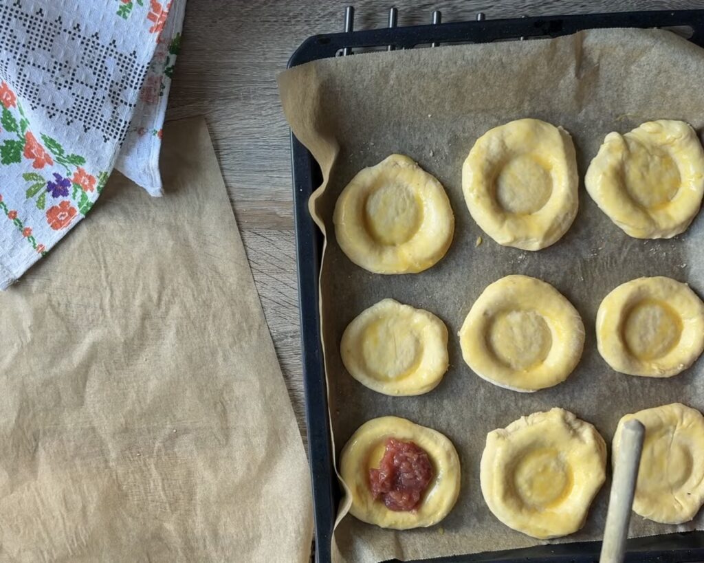 A baking tray with unbaked round pastries, including a partially filled Polish Apple Yeast Bun (Drożdż&oacute;wki) with jam, sits on parchment paper next to a cloth and a sheet of brown paper&mdash;an inviting start to an easy recipe.