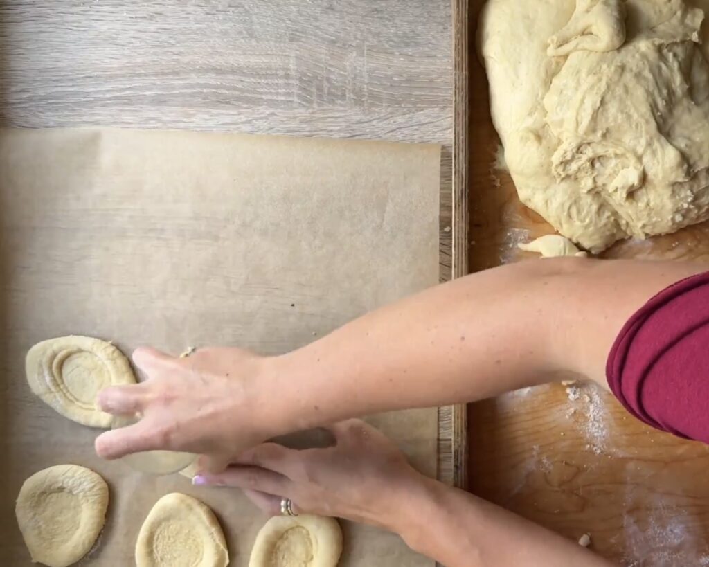 A person shapes dough rounds on parchment paper, preparing Polish apple yeast buns&mdash;drożdż&oacute;wki&mdash;with more dough and flour on the wooden surface nearby in this easy recipe.