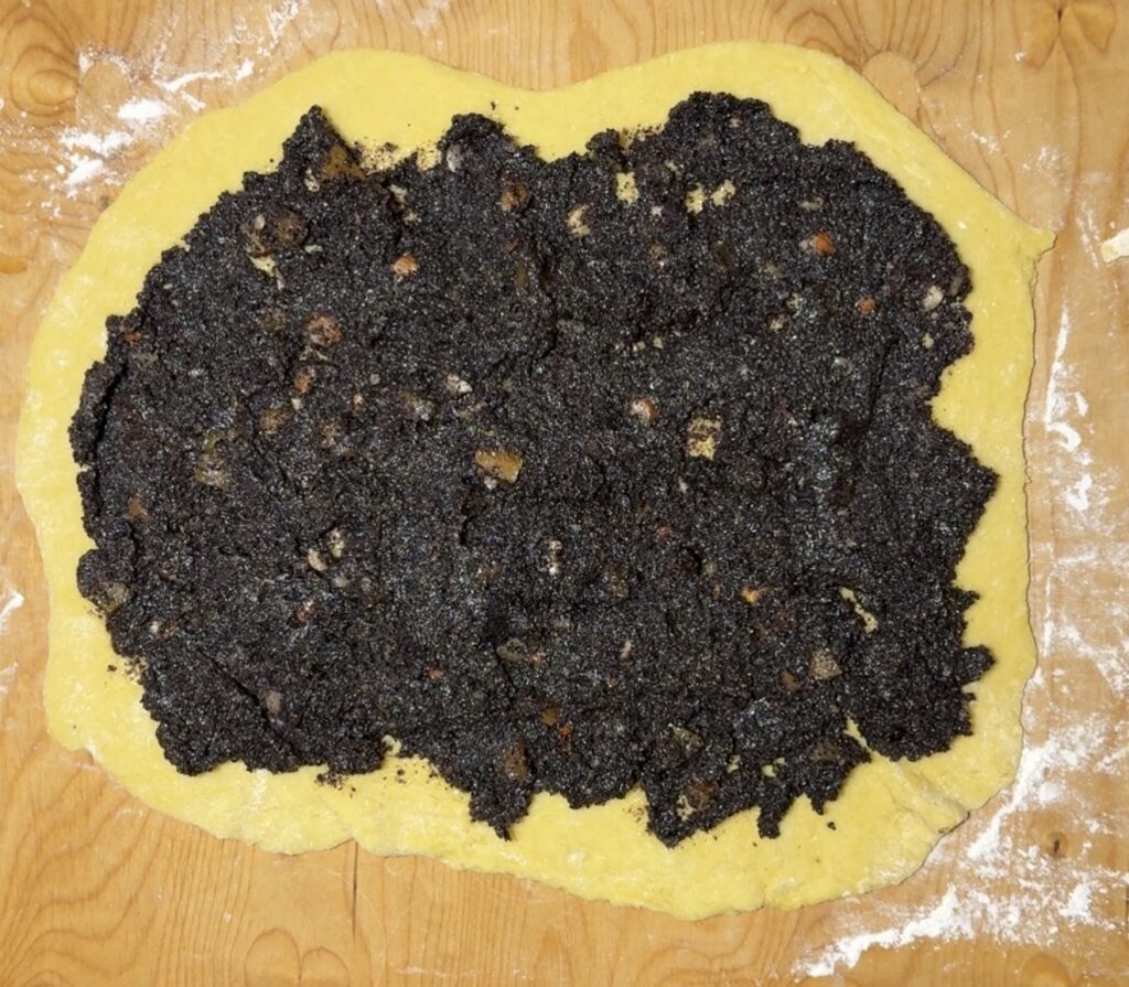 A sheet of dough topped with a dark poppy seed filling is spread out on a wooden surface dusted with flour, ready to be rolled into a delicious Makowiec or poppyseed twisted cake recipe.