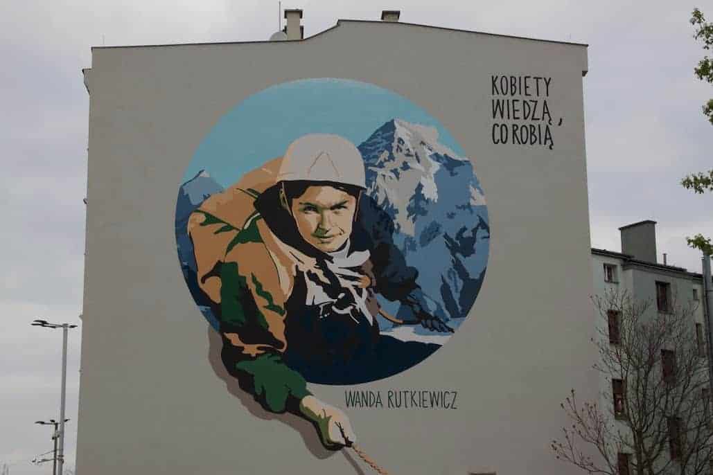 A mural of mountaineer Wanda Rutkiewicz climbing, set against a blue circle with mountains in Wroclaw; Polish text reads "Kobiety wiedzą, co robią" and her name appears below&mdash;a striking piece of Street Art Wroclaw is known for.