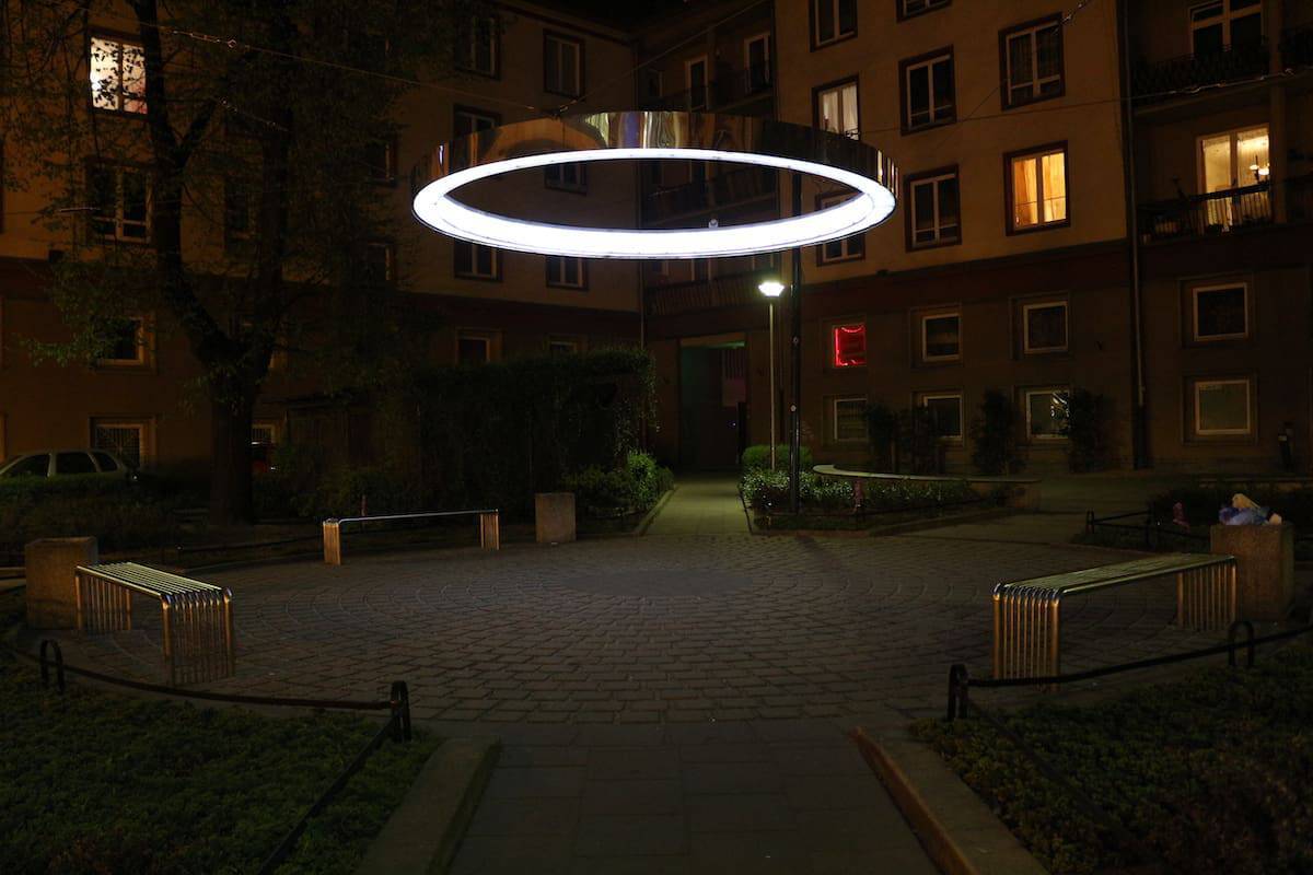 A circular, illuminated ring light hangs above a small courtyard with benches, surrounded by apartment buildings at night, echoing the creative flair seen in Wroclaw Street Art.