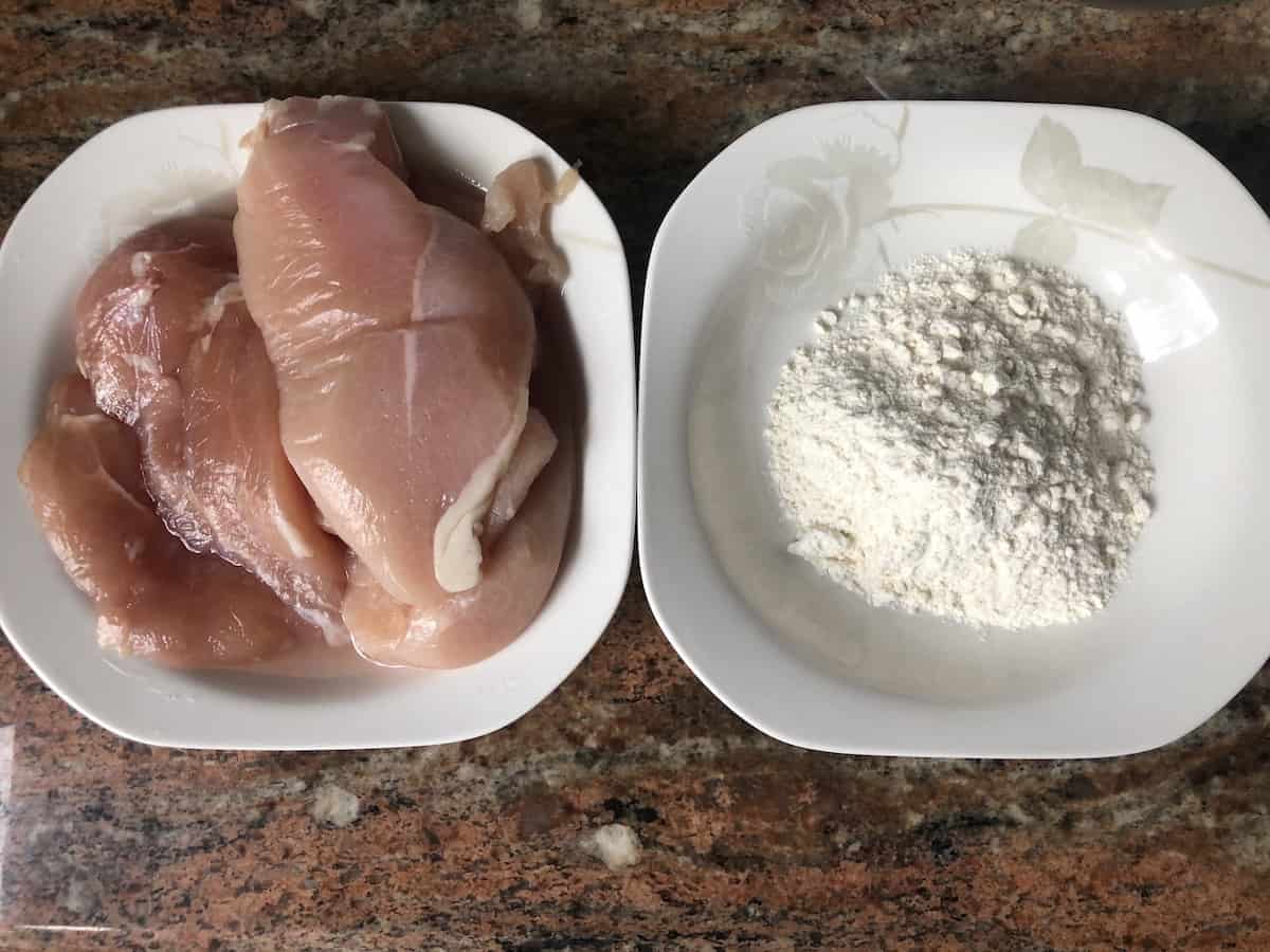 Uncooked chicken breasts and flour in a white plate.