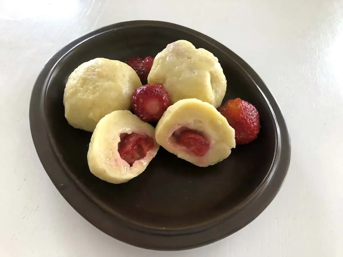 A brown plate with several dumplings, some whole and one cut open to reveal a strawberry filling, alongside whole strawberries.