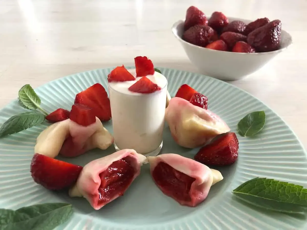 A light blue plate with strawberry-filled dumplings arranged around a small glass of white mousse, garnished with strawberry pieces and mint leaves. A bowl of whole strawberries in the background.