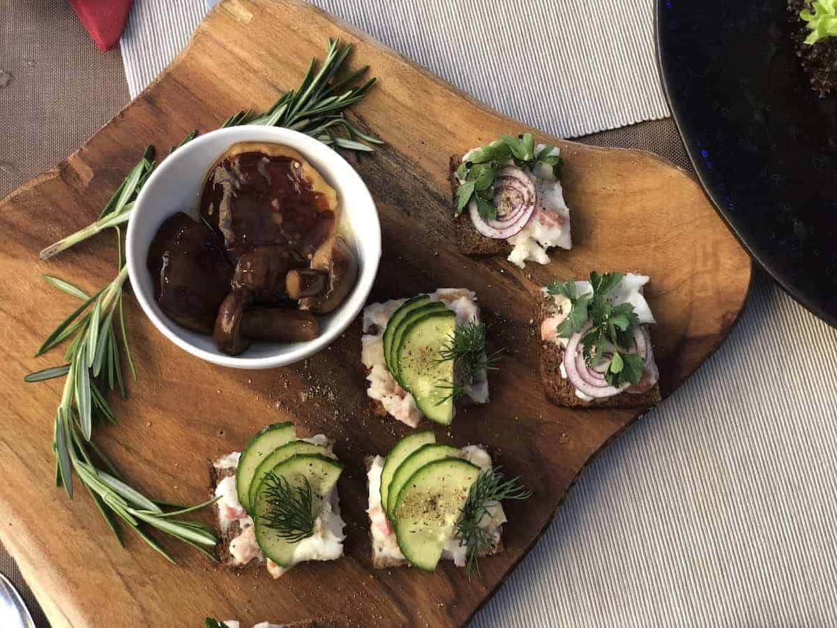 Wooden board with sliced bread topped with avocado, onions, and herbs; a small bowl of mushrooms in sauce sits on the board, garnished with rosemary sprigs. This delightful spread adds a Polish Twist to your 4th of July Feast.
