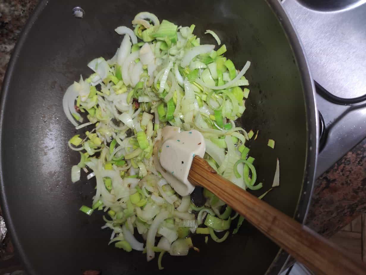 Sliced leeks and onions being saut&eacute;ed quickly in a pan with a wooden spatula, prepping for a delicious chickpea and sausage recipe.
