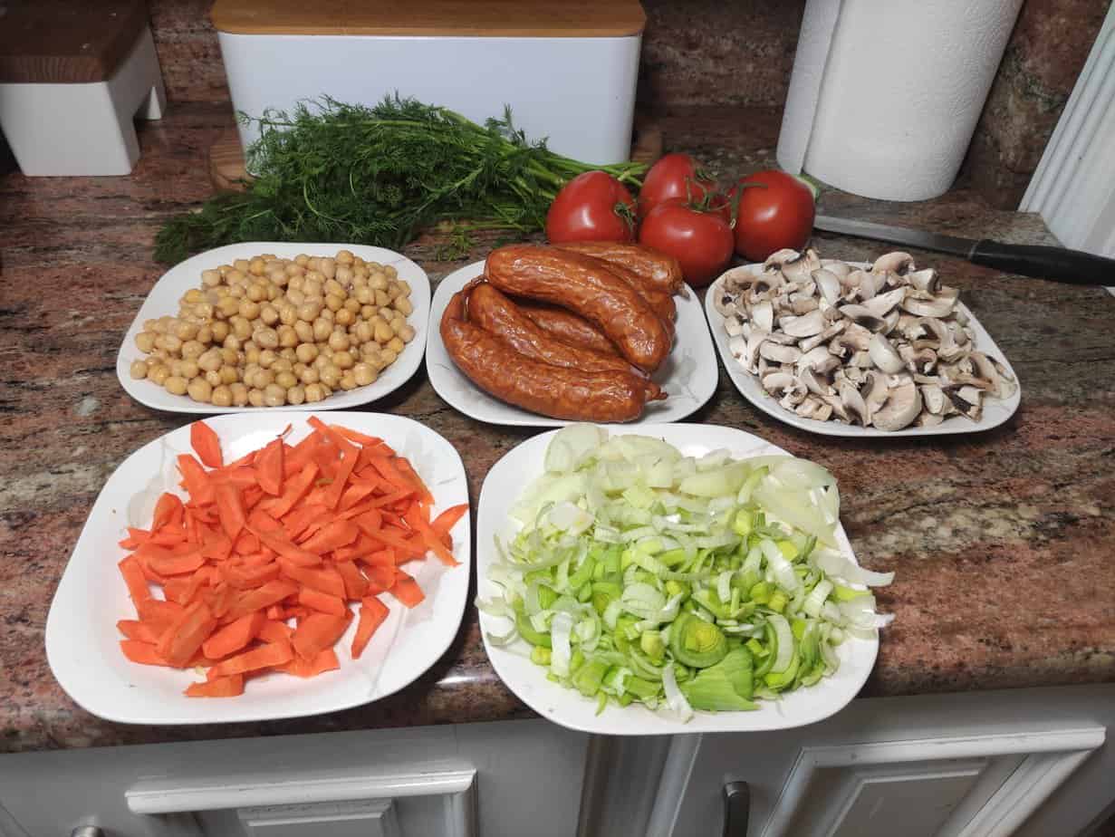 Assorted ingredients prepared for cooking, including chickpeas, sausages, mushrooms, carrots, leek, and tomatoes on a kitchen countertop for a delicious Chickpeas and Sausage
