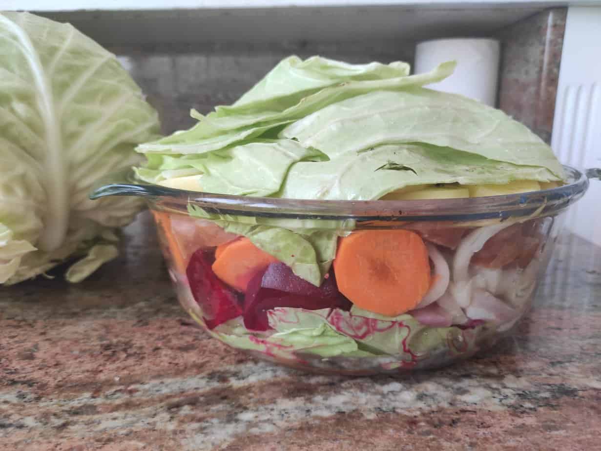 A glass bowl on a granite countertop filled with assorted chopped vegetables, including cabbage, carrots, and beets. A whole cabbage is partially visible on the left, reminiscent of ingredients for a traditional Polish Campfire Casserole (Prażonki).