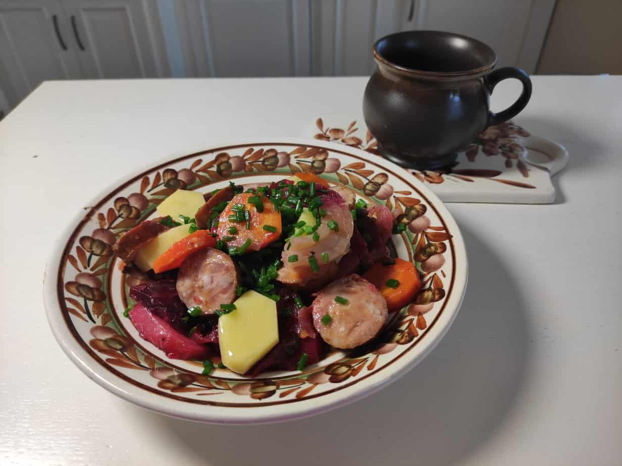 A bowl of sliced sausage, cheese, carrots, and leafy greens garnished with chopped chives resembles a modern twist on the traditional Polish Campfire Casserole. A brown ceramic mug sits on a coaster in the background.