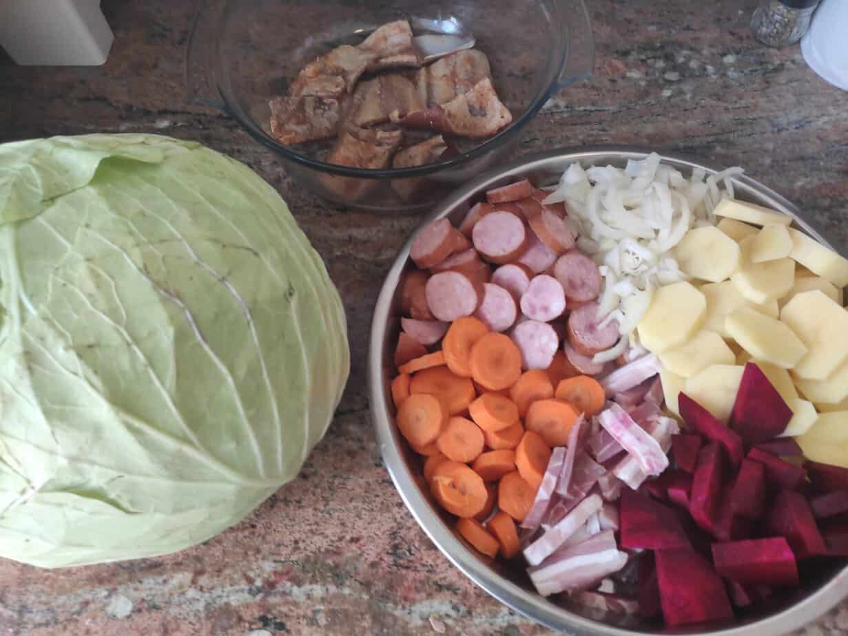 A whole cabbage, a bowl with cooked meat pieces, and a plate with sliced carrots, potatoes, onions, sausages, and beets are arranged on a countertop&mdash;perfect ingredients for an authentic Polish Campfire Casserole Recipe.