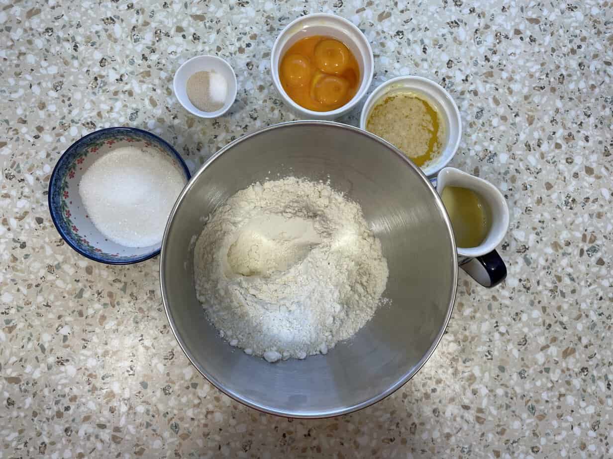 Top-down view of a kitchen counter with baking ingredients for Polish Nut Roll, including flour in a mixing bowl, eggs, sugar, oil, and butter, arranged neatly for preparation.