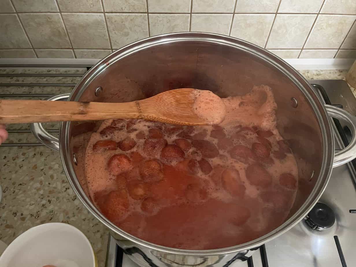 A wooden spoon stirring a pot of boiling strawberries on a gas stove, foam forming on top of the liquid. A small white bowl is partially visible in the bottom left corner. This scene is reminiscent of preparing traditional Dżem Truskawkowy, or Polish Strawberry Jam.