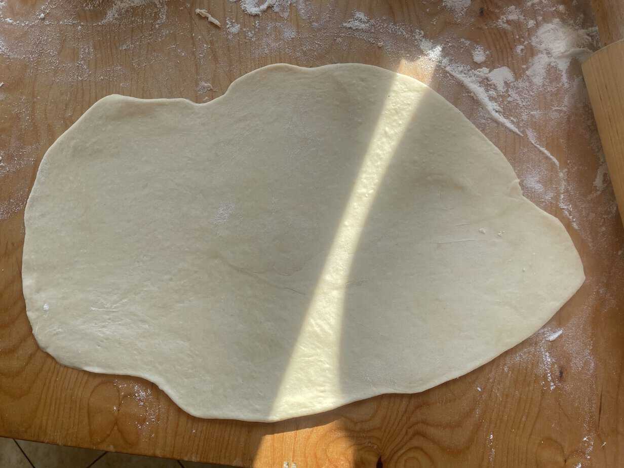 A sheet of pizza dough is rolled out on a floured wooden surface, with a partially illuminated area from sunlight, reminiscent of preparing Polish Sweet Cheese Twists like Zawijane Drożdż&oacute;wki.