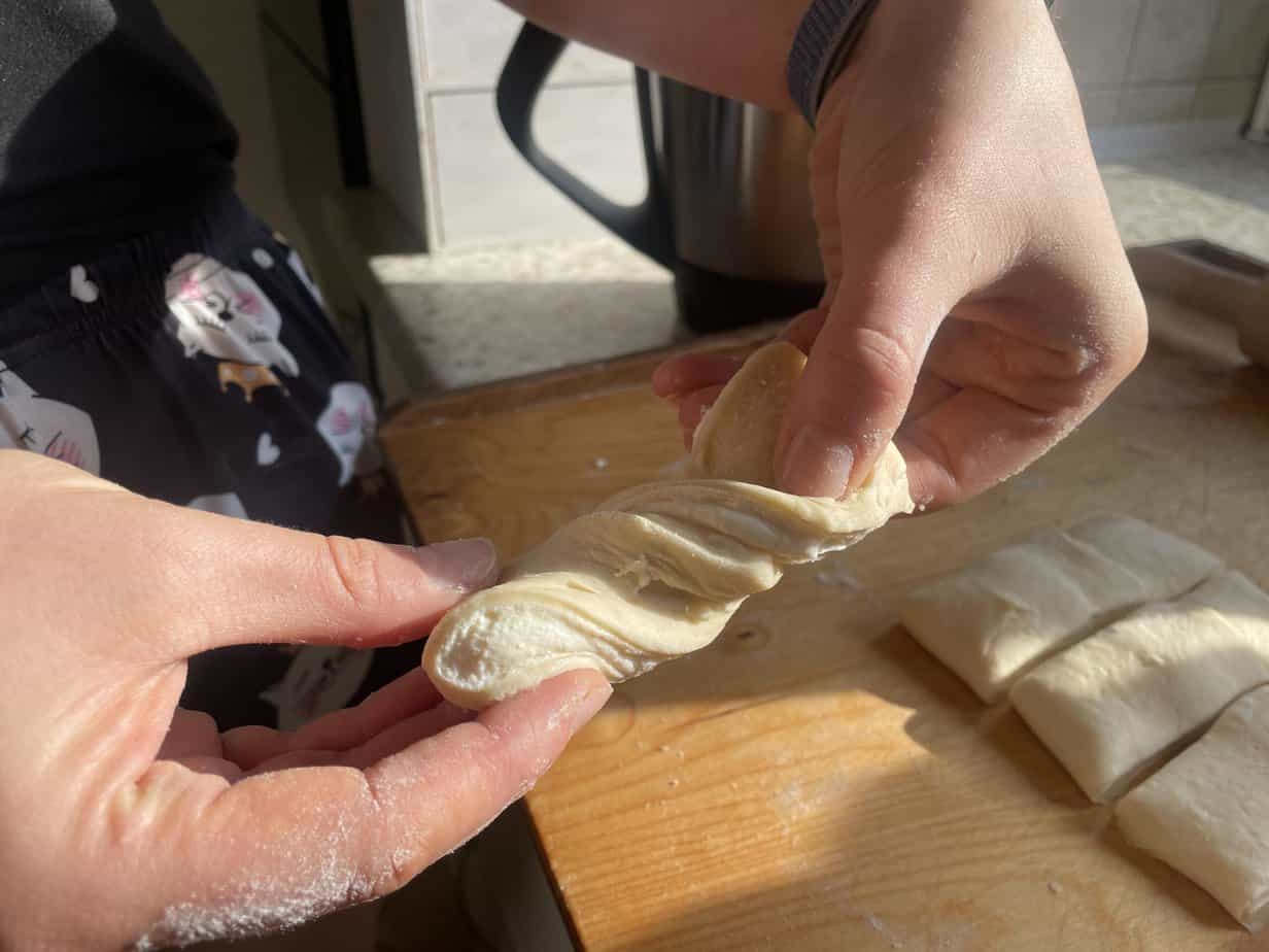 Hands twisting a piece of dough in a kitchen setting, preparing Zawijane Drożdż&oacute;wki with Serem, with cut dough portions nearby on a wooden surface.