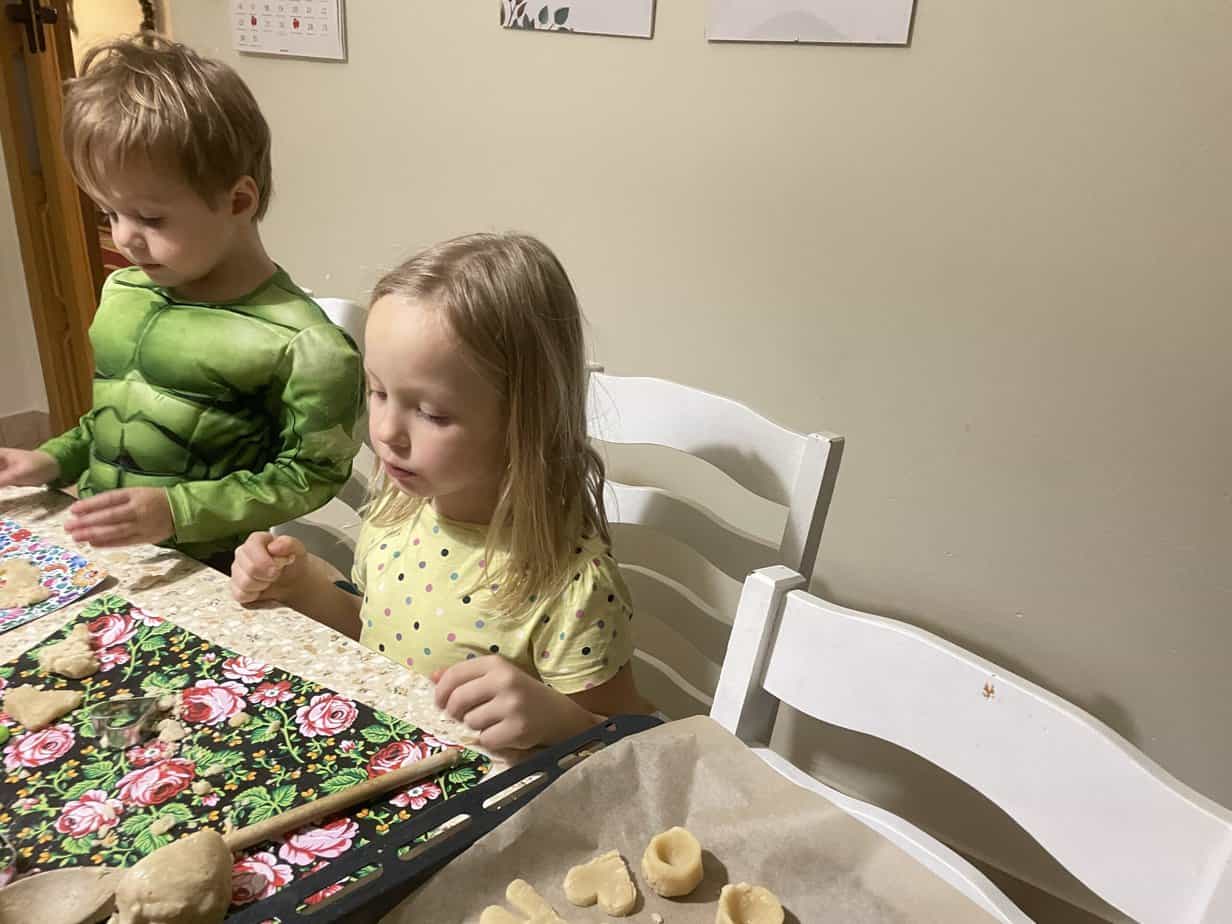 Two children sit at a table making Polish Marzipan Cookies. One wears a green superhero costume, while the other sports a yellow polka dot shirt. Cookie dough and freshly baked Marcepan Kr&oacute;lewiecki are spread across the table, turning their recipe adventure into a playful baking session.