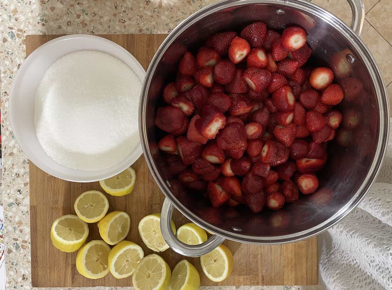 A large metal pot filled with halved strawberries sits on a wooden cutting board next to a bowl of sugar and sliced lemons, ready to be transformed into delicious Strawberry Jam. This traditional Polish Strawberry Jam Recipe, or Dżem Truskawkowy, is perfect for preserving the sweet taste of summer.