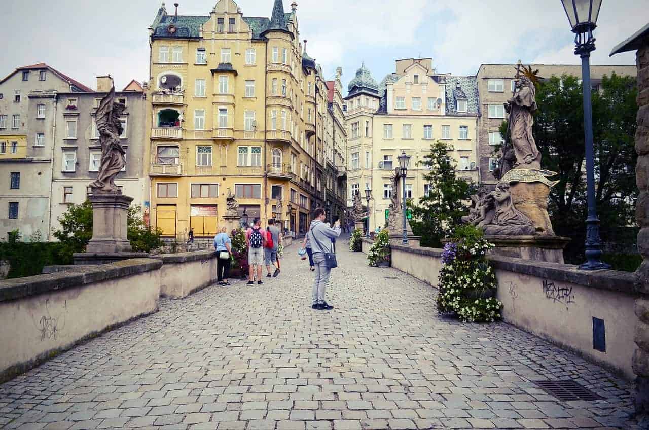 People walk across a cobblestone bridge with statues and flower planters, surrounded by historic European buildings under a partly cloudy sky&mdash;one of Dolnośląskie&rsquo;s best places to visit for unforgettable Poland travel experiences.