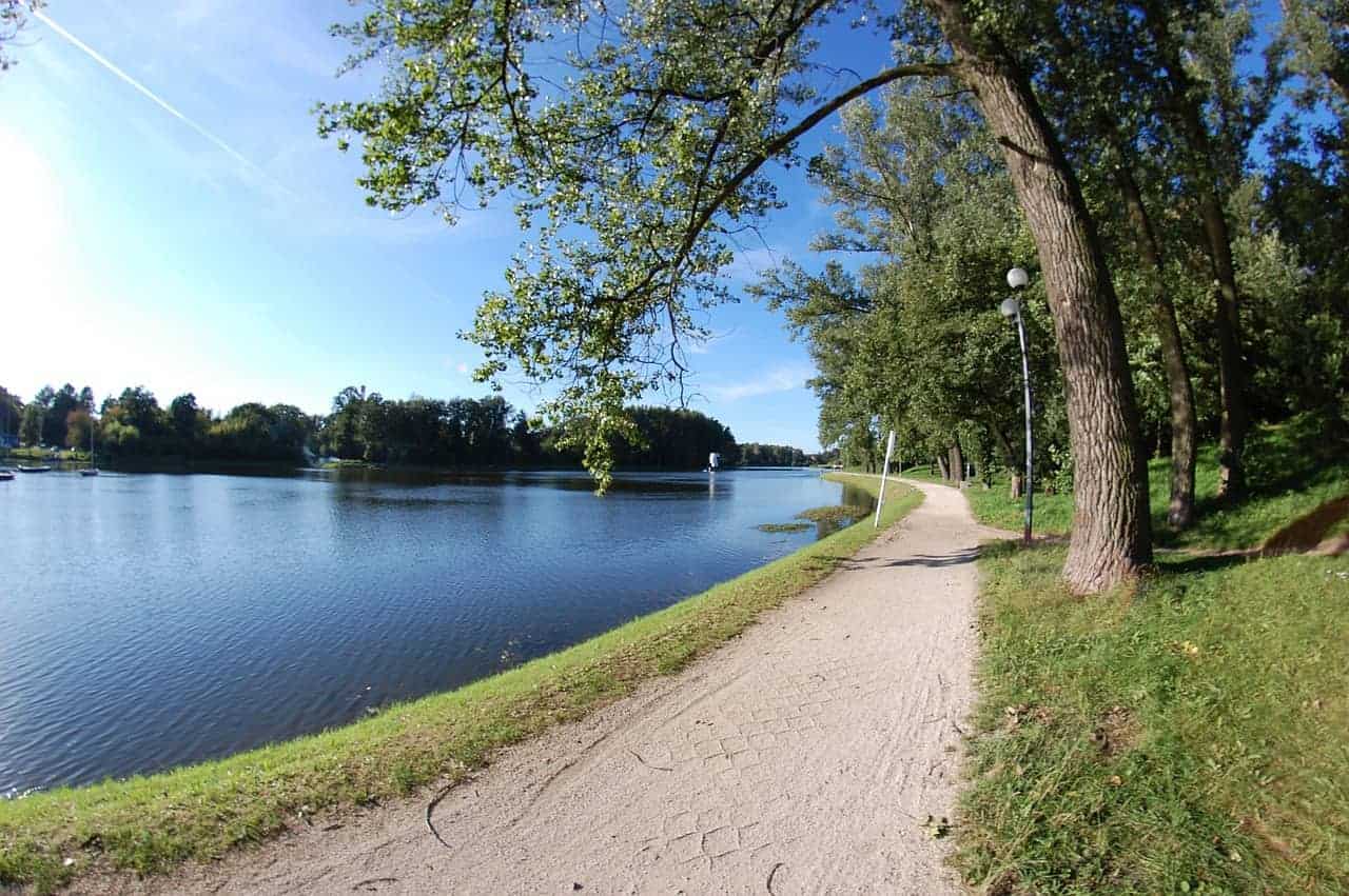 A gravel path runs alongside a calm river bordered by grass and trees on a clear, sunny day.