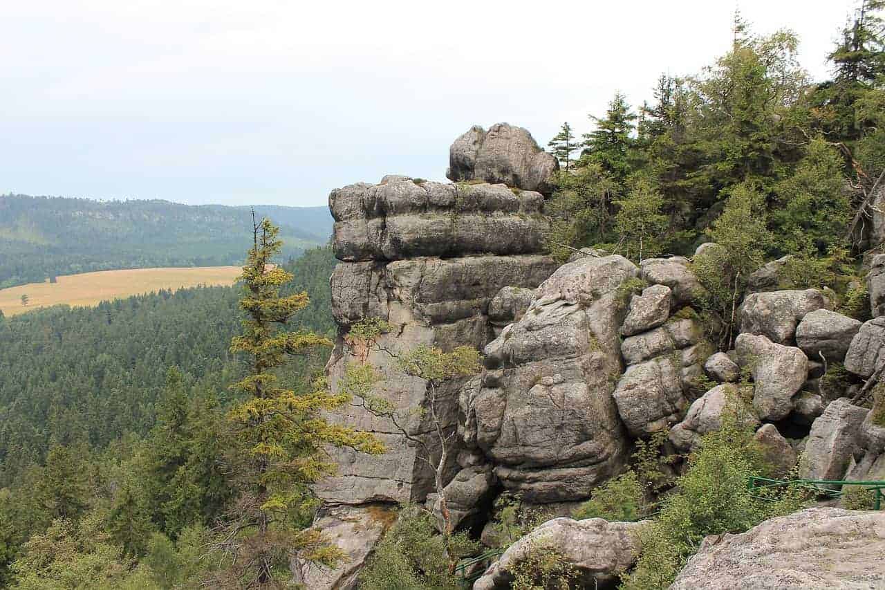 Large rock formations with layered stone surfaces rise above a forested landscape, with trees and distant hills visible under a cloudy sky&mdash;an iconic view when you visit Dolnośląskie, Poland.