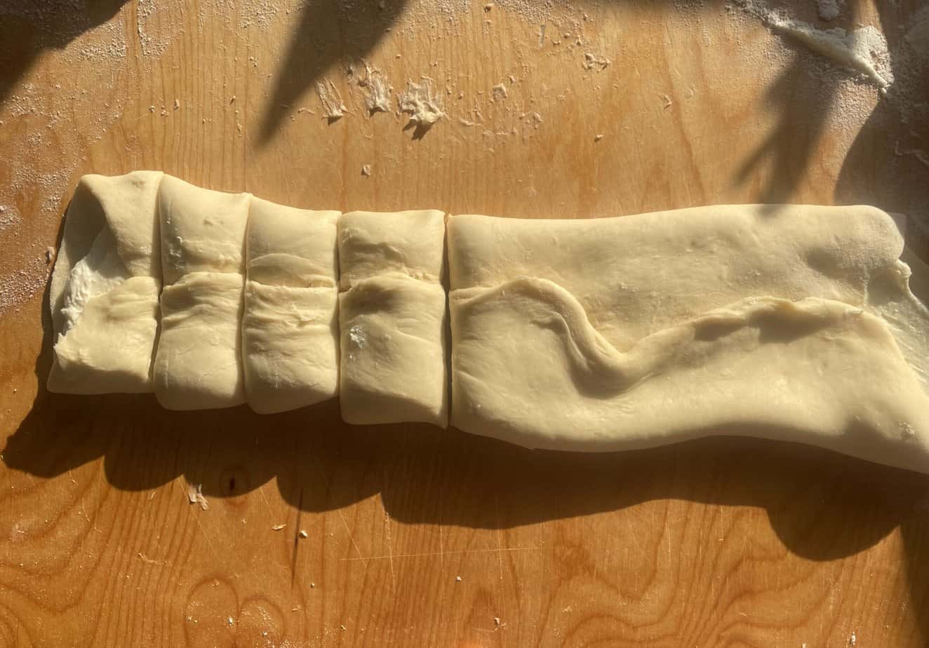 A rolled-out piece of dough is cut into several sections on a wooden surface, preparing the base for delicious Zawijane Drożdż&oacute;wki.