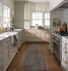 A bright, modern farmhouse-style kitchen with white shaker cabinets, brass hardware, marble countertops, and hardwood floors.