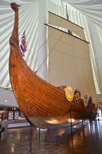 Full-scale replica of a Viking ship on display at the Hjemkomst Center in Moorhead, MN, under a bright vaulted ceiling with a Norwegian flag at the stern.