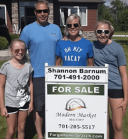 Happy family standing in front of a home for sale with a Modern Market REALTORS&reg; sign featuring Shannon Barnum&rsquo;s contact information.