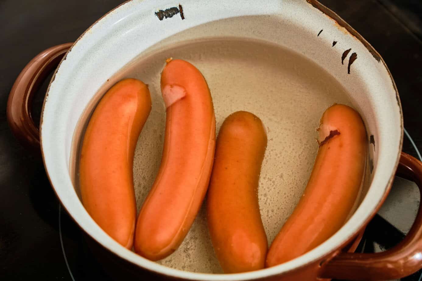 Three whole Polish sausages cooking in a beige ceramic pan on a stove.