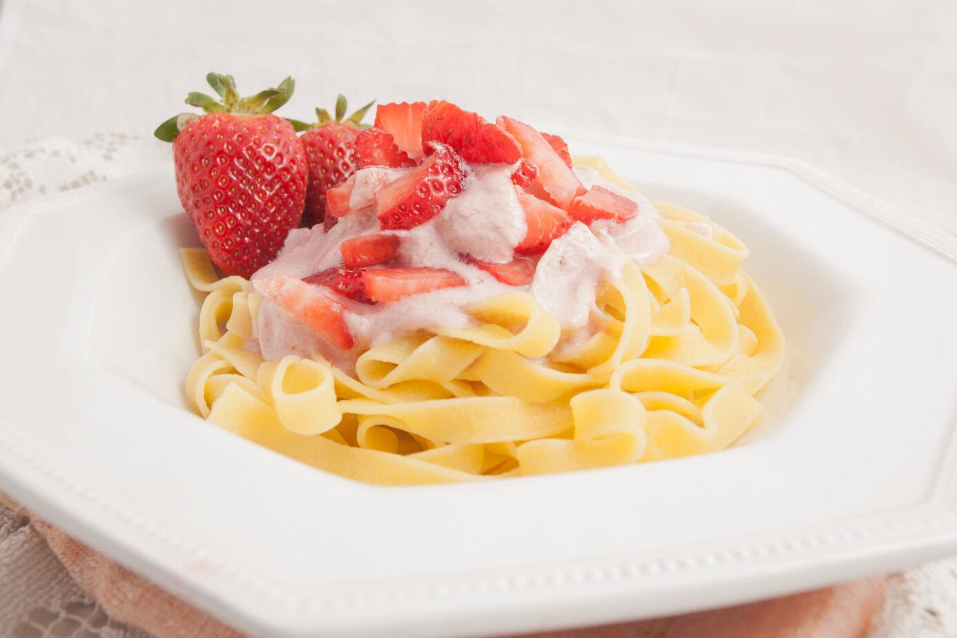 A white plate of tagliatelle pasta topped with sliced strawberries and strawberry cream sauce, garnished with whole strawberries.
