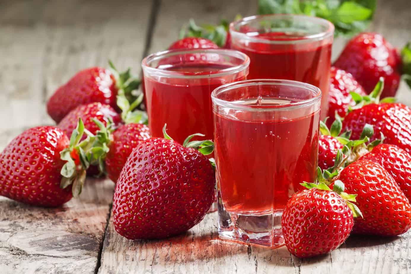Three glasses of strawberry juice are on a wooden table surrounded by fresh strawberries.