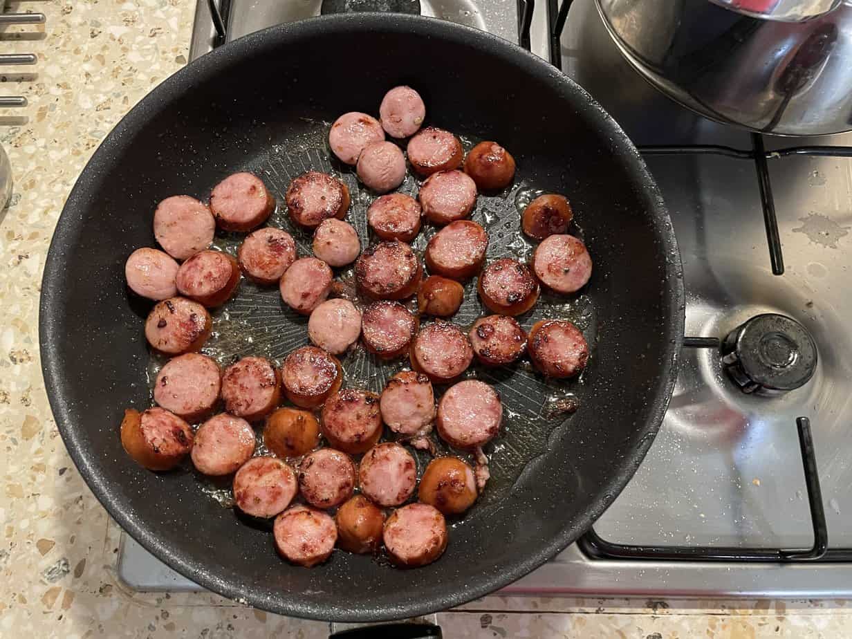 Polish sausages sliced and cooking in oil in a frying pan on a stove.