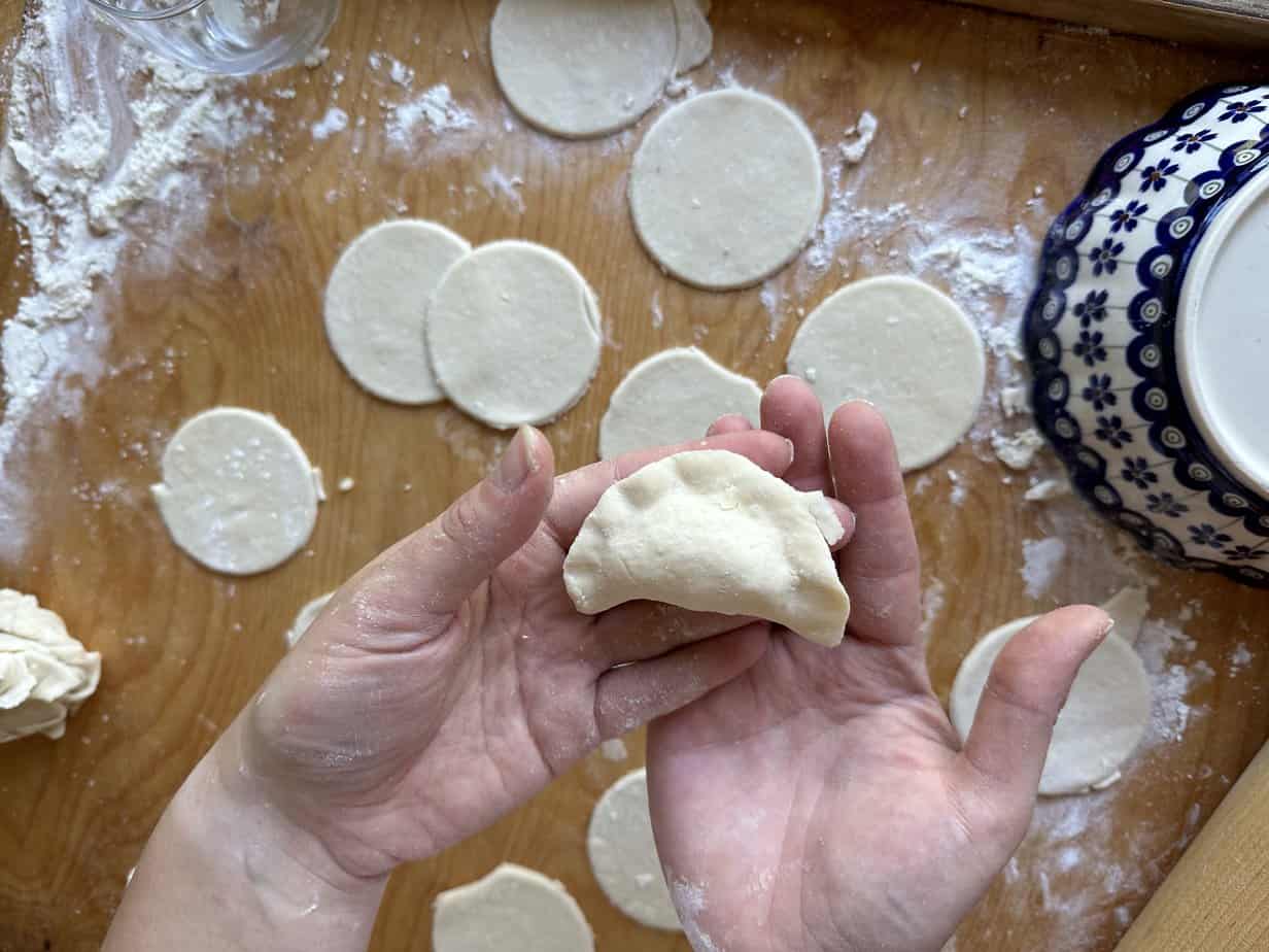 Hands shaping a dumpling with circular dough pieces laid out on a floured wooden surface, reminiscent of the age-old question: Where Did Pierogi Originate From?