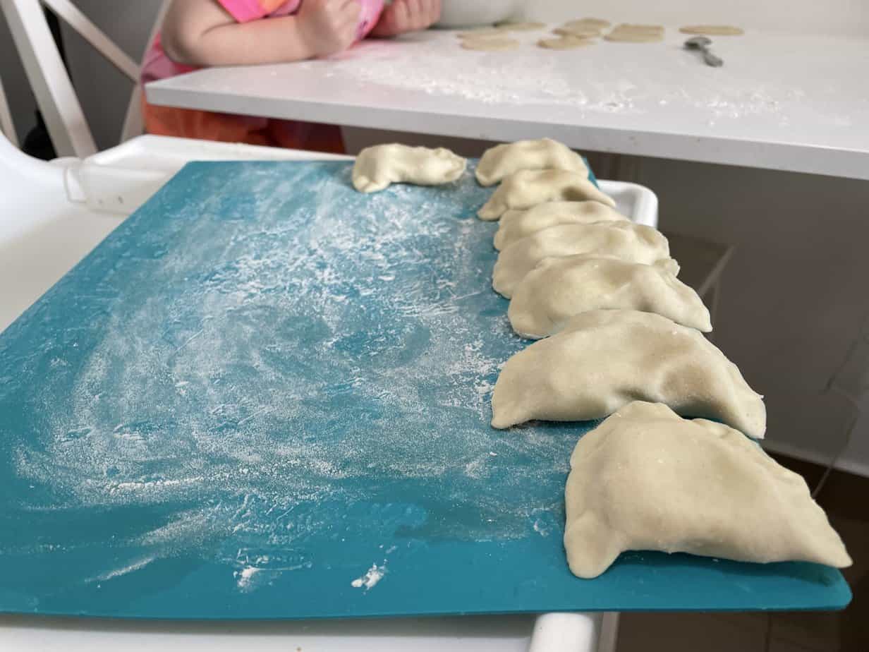 A row of uncooked dumplings is arranged on a blue, flour-dusted surface, with a child partially visible in the background, preparing more dumplings at a table. One can't help but wonder where did pierogi originate from as the scene evokes a sense of timeless tradition.