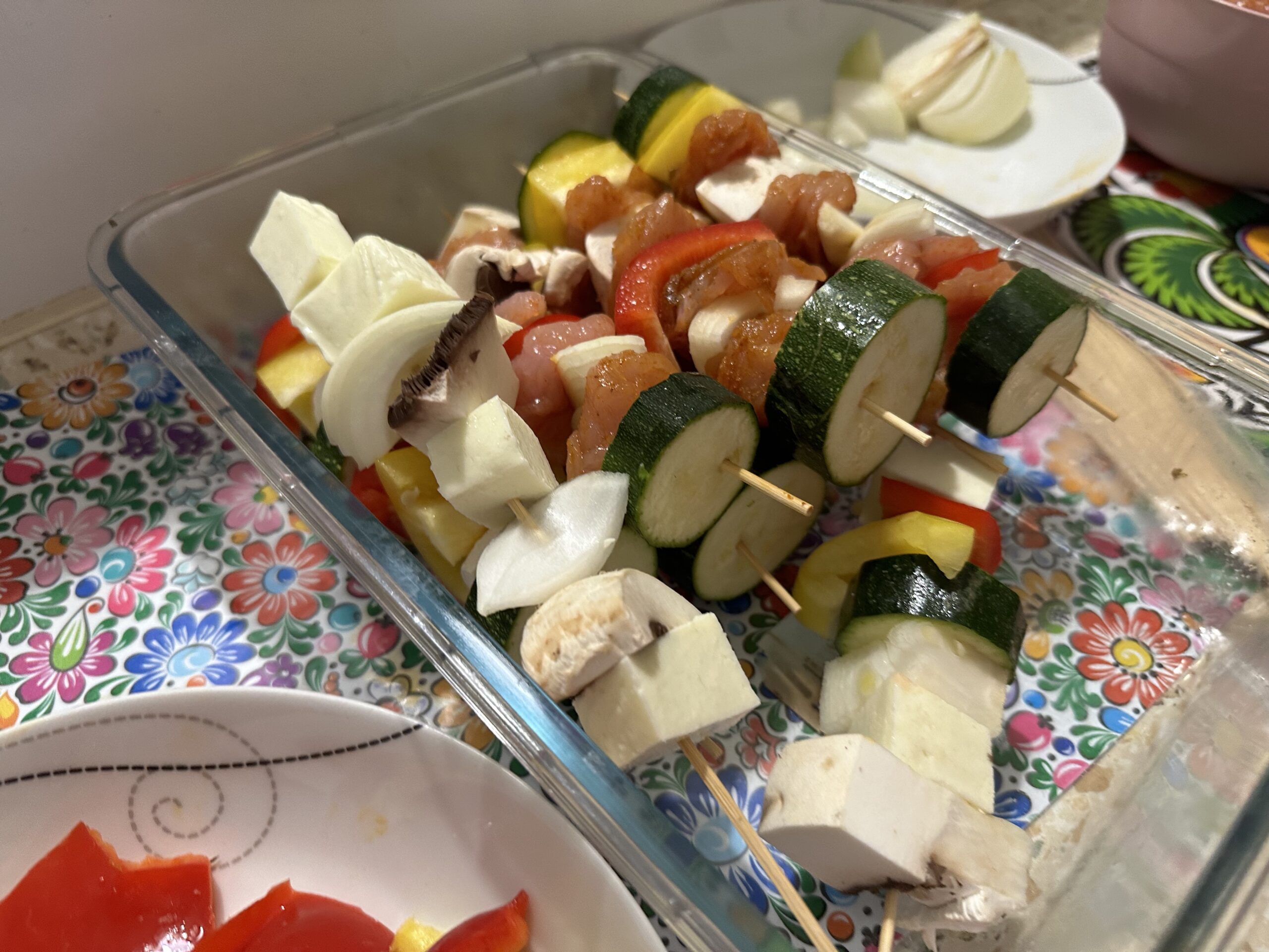 A tray of freshly prepared szaszłyki with zucchini, tomatoes, mushrooms, onions, and bell peppers on a colorful floral plate.