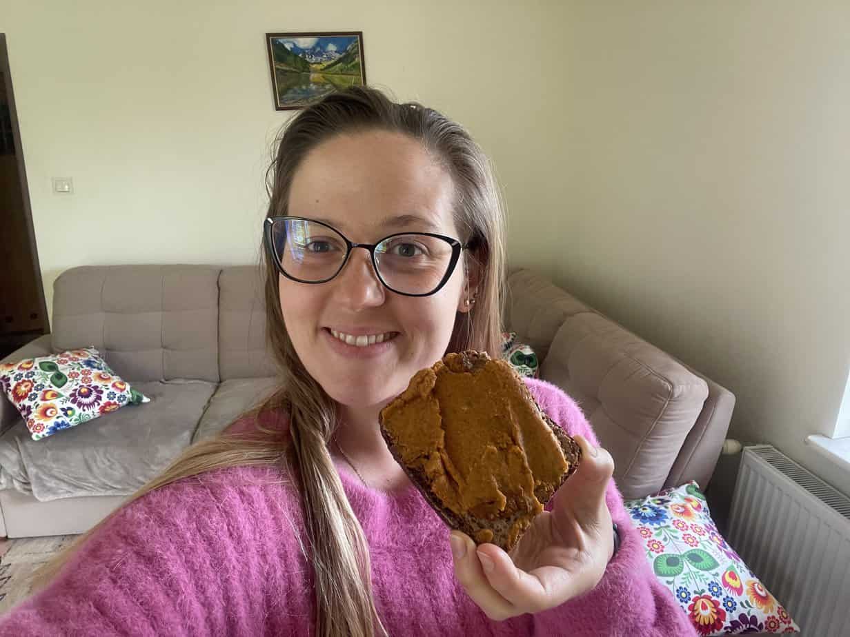 A woman wearing glasses and a pink sweater holds up a piece of bread with vegan pate. She is smiling and standing in a living room with a beige couch and colorful pillows.