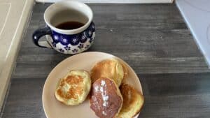 A floral-patterned mug filled with dark liquid sits beside a plate of four round, golden-brown Polish apple pancakes on a gray countertop.