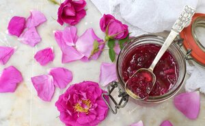 A jar of Konfitura R&oacute;żana with a silver spoon, surrounded by scattered pink rose petals on a marble surface, and a white lace cloth nearby.
