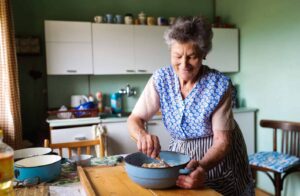 Elderly woman in a blue apron smiles while mixing ingredients in a bowl at a kitchen table, surrounded by baking supplies to celebrate Mother's Day.