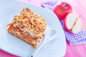 A slice of Polish apple cake with meringue on a plate with a spoon, accompanied by a whole apple and a half-cut apple in the background.