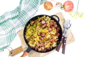 A cast iron skillet filled with cooked cabbage and sliced traditional Polish kielbasa on a wooden board, accompanied by a green checkered cloth, onion, garlic, apple, and cutlery on the side.