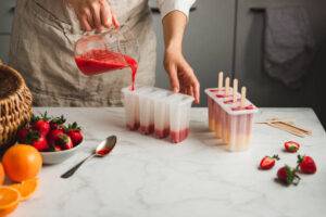 Fun-Easy-Homemade-Ice-Lollies-Popsicles-—-The-Perfect-Summer-Treat-image-1-person-pouring-fruit-puree-into-ice-lolly-moulds-with-fresh-fruit-scattered-Frosted-Fusions