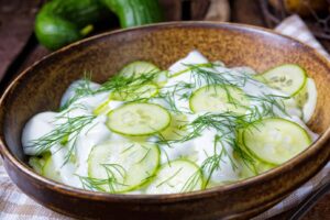A wooden bowl filled with Polish cucumber salad&mdash;crisp sliced cucumbers in a creamy white dressing, garnished with fresh dill, with whole cucumbers visible in the background.