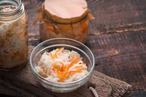 A glass bowl filled with shredded sauerkraut and carrots sits on a wooden board, showcasing a classic kapusta kiszona recipe; beside it are two jars of sauerkraut, one open and one closed with a kraft paper lid.