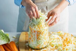 A person wearing an apron packs shredded cabbage and carrots into a glass jar, reminiscent of authentic Polish recipes with sauerkraut, with additional shredded vegetables and whole carrots on a wooden cutting board.