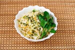 A white bowl filled with Polish sauerkraut salad, garnished with fresh parsley, placed on a woven mat.