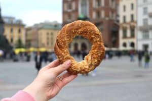 A hand holding a round, sesame-topped bread with a bite taken out, in focus against a blurred city square background.