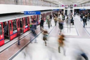 People travel around Poland as they move through a busy subway station, with red trains stopped at the platform and signs above indicating directions, including one reading "Kabaty.